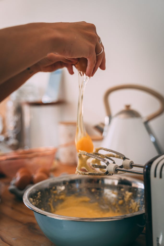 A hand cracks an egg into a mixing bowl amidst baking preparation in a cozy kitchen environment.