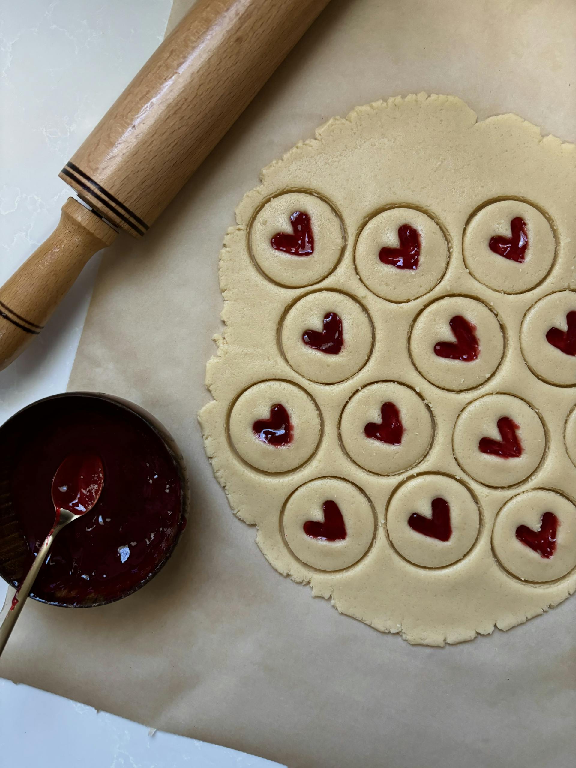 Heart-shaped cookies with jam being prepared on parchment paper.