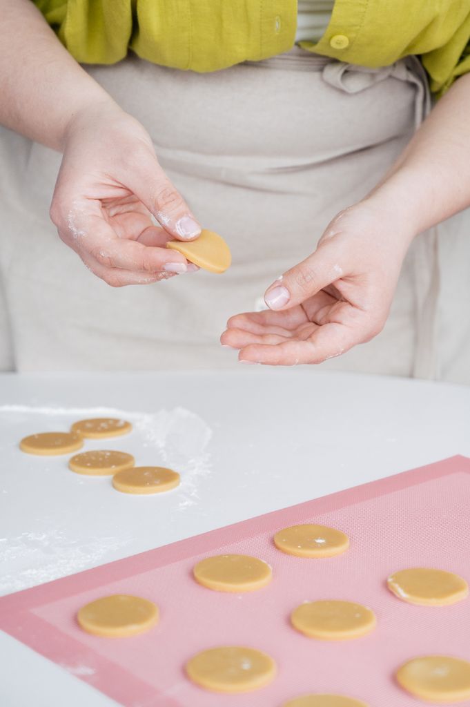 Crop unrecognizable female confectioner in apron putting raw homemade cookies on baking paper while standing at white table