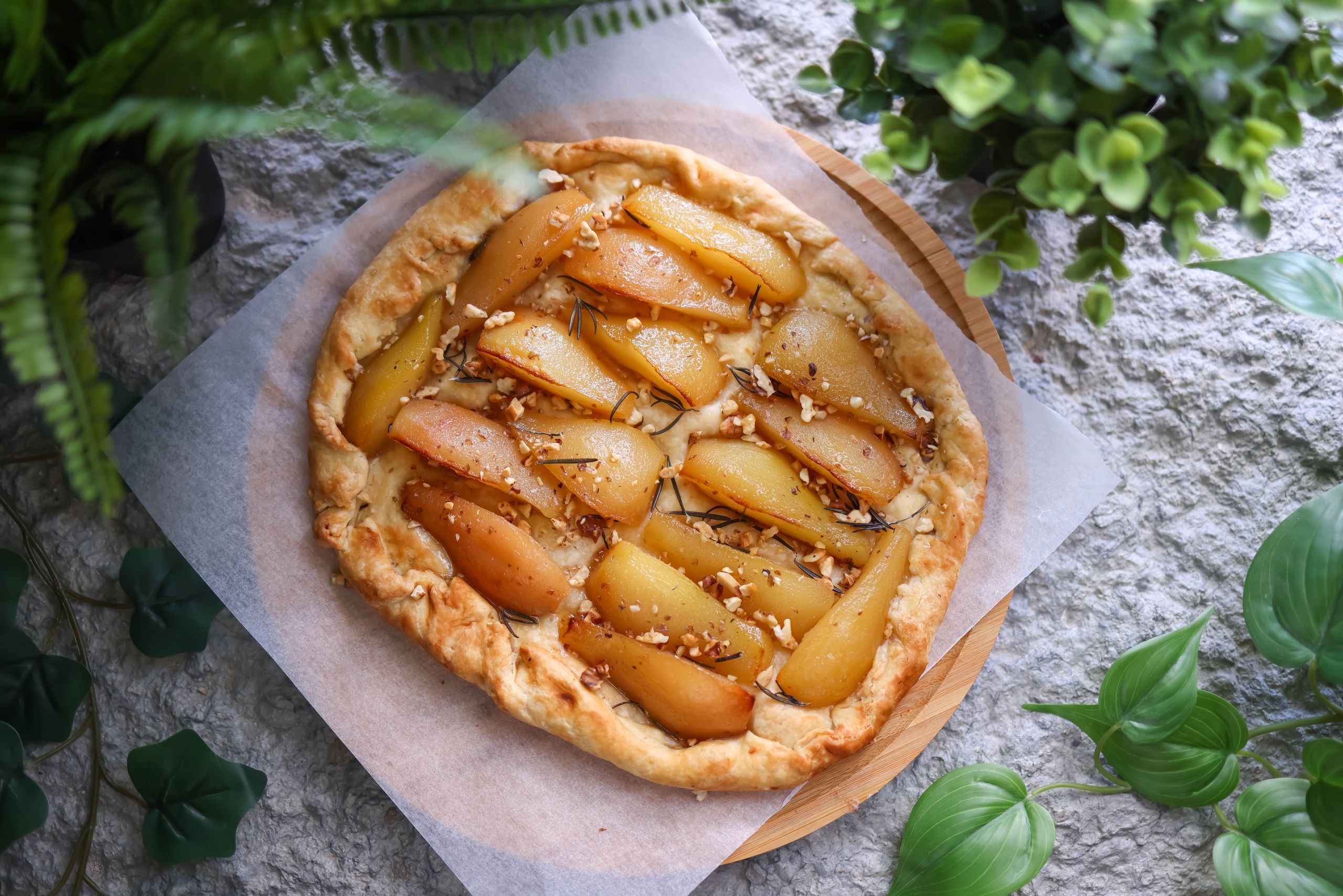 Homemade pear tart with nuts and herbs on a rustic stone background surrounded by greenery.
